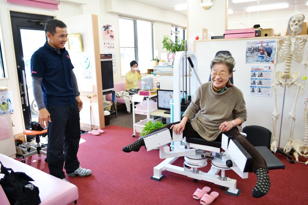 しいな整骨院の院内の様子。椎名先生と、インナーサイに取り組む笑顔の高齢女性の構図。